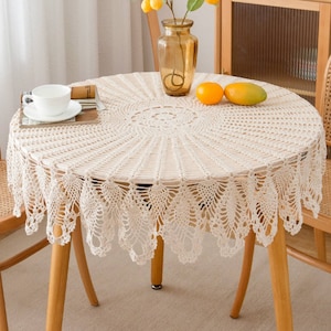 May include: A cream-coloured, round crochet tablecloth with a scalloped edge, covering a wooden table. A vase with yellow flowers, a cup and saucer, and fruit are on the table. The table has light brown wooden legs.
