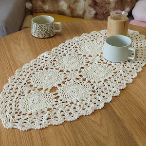 Peut inclure: Un chemin de table ovale en crochet blanc avec un motif de dentelle délicat. Le chemin de table a un bord festonné. Il est posé sur une table en bois.