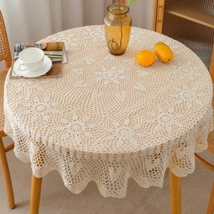 Peut inclure: Une nappe ronde en crochet de couleur crème avec un motif floral délicat. La nappe est drapée sur une table en bois avec deux chaises.