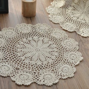 Puede incluir: Dos manteles individuales de ganchillo beige con un delicado patrón floral. Los manteles individuales son redondos y tienen un borde festoneado.