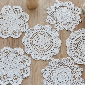 Può includere: Sei tovagliette all'uncinetto bianche con motivi intricati, disposte su una superficie di legno.