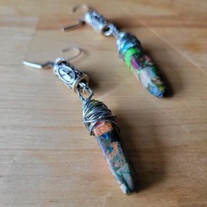 May include: A pair of dangle earrings with silver wire wrapped around a long, pointed, multi-colored stone. The stones are a mix of green, blue, orange, and brown.