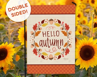 May include: A double-sided garden flag with a fall theme. The flag features the words "HELLO autumn" surrounded by a wreath of leaves and acorns. The top and bottom borders have a plaid design. The background is a field of sunflowers.