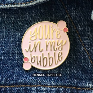 May include: A pink enamel pin with a gold outline and the text "You're in my bubble" in gold lettering. The pin has three small pink circles around the text. The pin is on a blue denim jacket. The text "HENNEL PAPER CO." is below the pin.