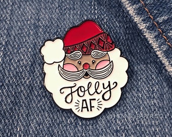 May include: A white enamel pin with a cartoon Santa Claus face and the text "Folly AF" in black cursive lettering. The Santa Claus has a red hat with a white trim and a white beard.