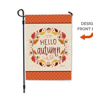May include: A garden flag with a fall theme. The flag features the words "HELLO autumn" in a decorative font, surrounded by a wreath of leaves and acorns. The top and bottom borders have an orange plaid pattern. The flag is attached to a black metal pole.
