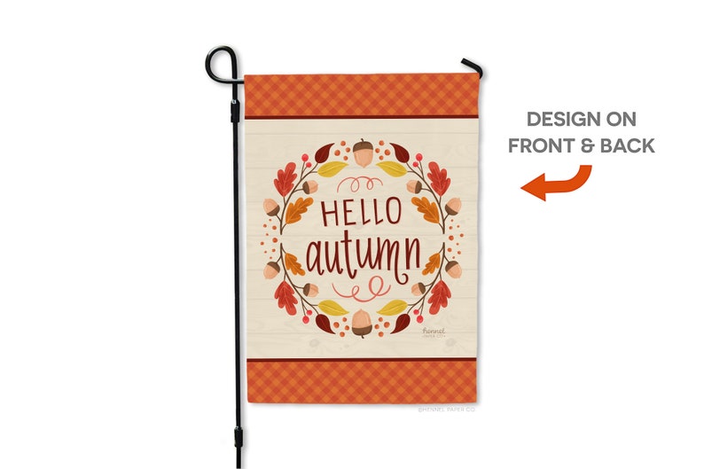 May include: A garden flag with a fall theme. The flag features the words "HELLO autumn" in a decorative font, surrounded by a wreath of leaves and acorns. The top and bottom borders have an orange plaid pattern. The flag is attached to a black metal pole.