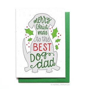 May include: A white card with a gray dog illustration and the text "merry Christ mas to the BEST Dog dad". The dog is surrounded by green holly leaves with red berries.