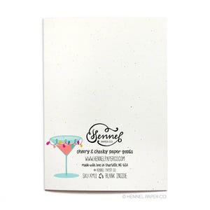 May include: Back of a blank greeting card with a light blue martini glass with colorful lights around the rim. The card is made by Sennel Paper Co. and has the text "cheery & cheeky paper goods" and "www.hennelpaperco.com".