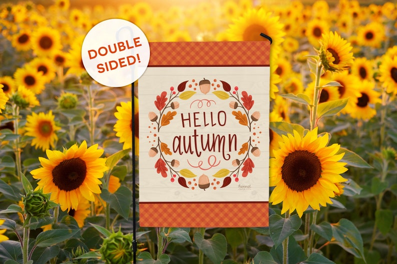 May include: A double-sided garden flag with a fall theme. The flag features the words "HELLO autumn" surrounded by a wreath of leaves and acorns. The top and bottom borders have a plaid design. The background is a field of sunflowers.