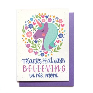 Puede incluir: Una tarjeta con un unicornio morado con una crin azul y una corona de flores a su alrededor. La tarjeta dice "Gracias por siempre creer en m&iacute;, mam&aacute;."