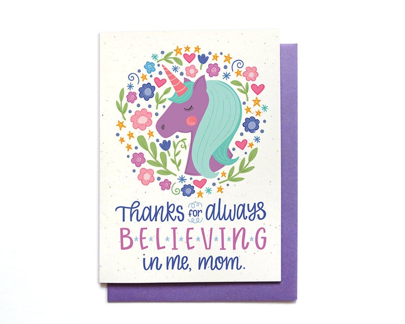 Puede incluir: Una tarjeta con un unicornio morado con una crin azul y una corona de flores a su alrededor. La tarjeta dice "Gracias por siempre creer en m&iacute;, mam&aacute;."