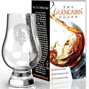May include: A clear glass Glencairn whisky glass with a wide base and a rounded bowl. The glass has a logo etched on the side that reads "Macdonald per Terras". The glass is sitting on a reflective surface. The background shows a box with the words "The Glencairn Glass" printed on it.