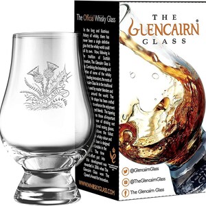Puede incluir: Una copa de whisky Glencairn transparente con un emblema de cardo y la palabra "SCOTLAND" grabada en la parte delantera. La copa se muestra junto a su embalaje, que presenta el texto "The Glencairn Glass" y una salpicadura de l&iacute;quido &aacute;mbar.
