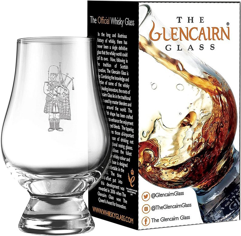 Puede incluir: Un vaso de whisky Glencairn transparente con una imagen grabada de un gaitero. El vaso se muestra junto a su embalaje, que presenta el texto "The Glencairn Glass" y una salpicadura de l&iacute;quido &aacute;mbar.