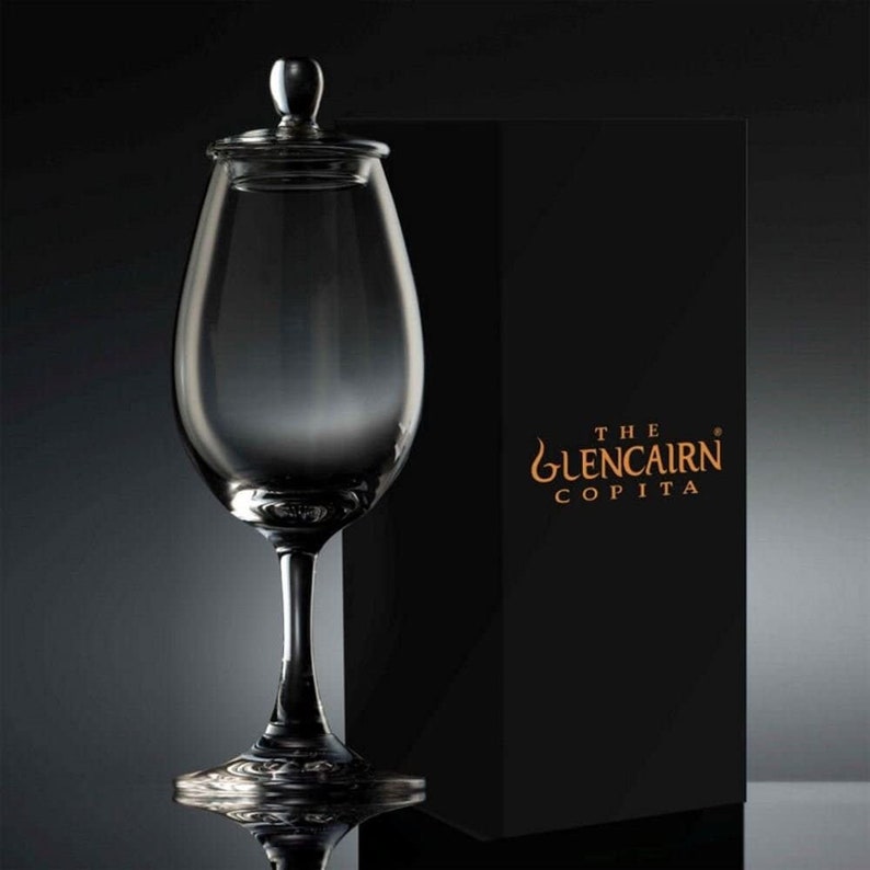 Puede incluir: Una copa copita de cristal transparente con tapa, presentada frente a una caja negra. La copa tiene un tallo largo y un cuenco ancho. La caja negra tiene la inscripci&oacute;n dorada "The Glencairn Copita".