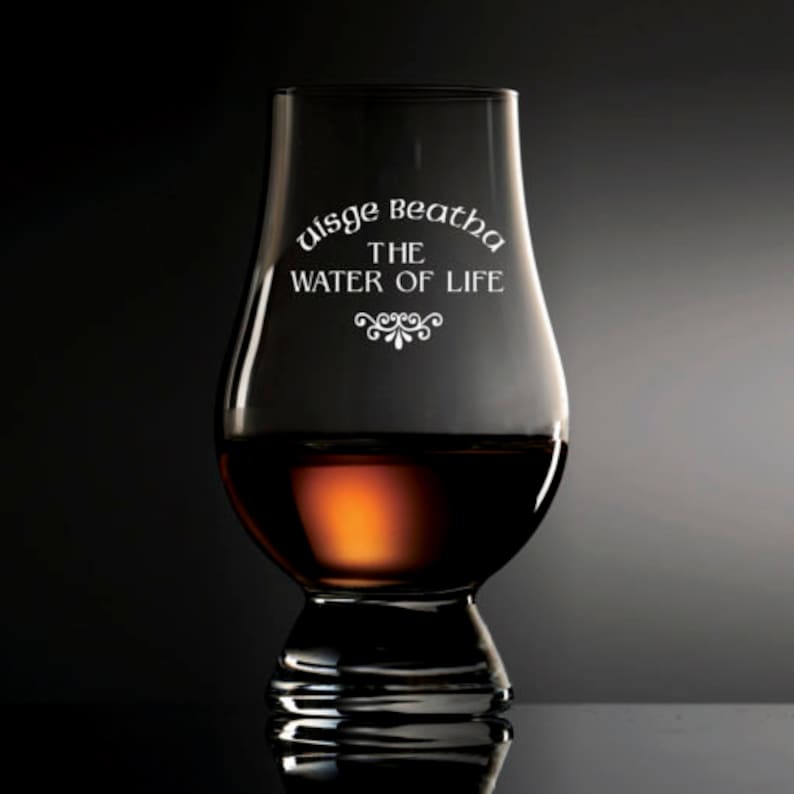 Puede incluir: Una copa de whisky transparente, en forma de tulip&aacute;n, llena de l&iacute;quido &aacute;mbar. La copa tiene el texto blanco "Uisge Beatha THE WATER OF LIFE". La copa est&aacute; sobre una superficie oscura.