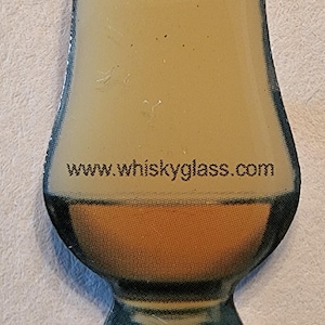 Puede incluir: Un objeto decorativo en forma de vaso con un diseño de whisky. La imagen presenta un vaso con una parte superior beige claro, un centro naranja y una base oscura. El texto "www.whiskyglass.com" está impreso en el vaso.