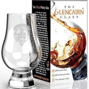 May include: A clear glass Glencairn whisky glass with a wide base and a rounded bowl. The glass has an etched design of a Scottish clan crest with the text "Touch Not This Cat Bot A While". The glass is displayed on a white surface with a box behind it that reads "The Glencairn Glass".
