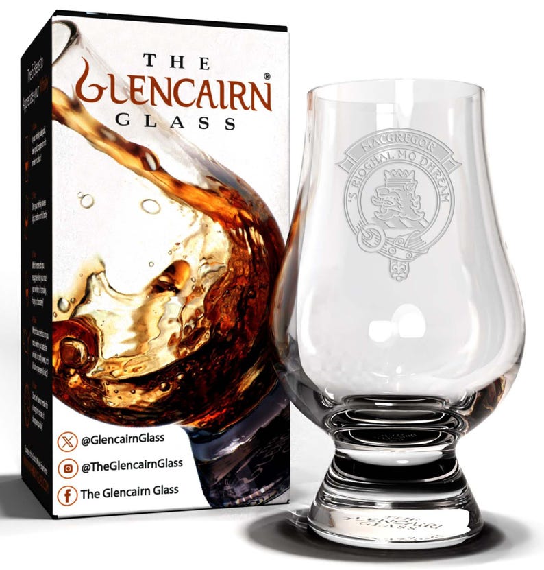 May include: A clear glass Glencairn whisky glass with an etched design of the MacGregor clan crest. The crest features a lion holding a sword and the text "MacGregor Ogha Mo Dhreah". The glass is sitting on a white surface.