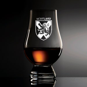 Puede incluir: Una copa de degustaci&oacute;n de whisky transparente con la palabra "SCOTLAND" y un escudo. La copa est&aacute; llena de l&iacute;quido color &aacute;mbar. El fondo es oscuro.