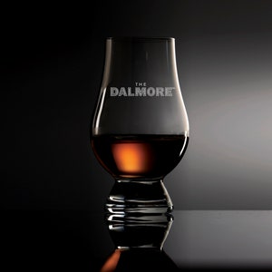 Puede incluir: Un vaso de vidrio transparente con una base ancha y una parte superior redondeada, lleno de líquido ámbar. El vaso tiene las palabras "THE DALMORE" grabadas en el lateral.