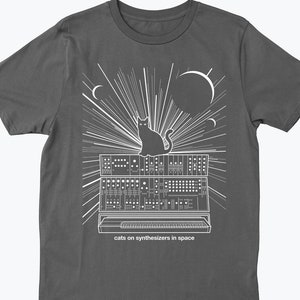 Puede incluir: Una camiseta gris oscuro con un diseño gráfico blanco de un gato sentado en un sintetizador con un fondo estrellado. El texto "cats on synthesizers in space" está impreso debajo del gráfico.