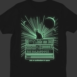 Puede incluir: Una camiseta negra con un diseño que brilla en la oscuridad de un gato sentado en un sintetizador en el espacio. El texto "cats on synthesizers in space" está impreso debajo del diseño.