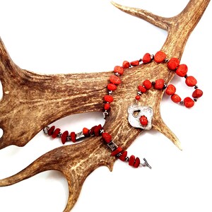 May include: A red coral bead necklace with a silver pendant, displayed on a brown antler against a white background. The necklace features a toggle clasp and small silver beads. The pendant has a unique, open design.
