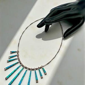May include: A silver necklace with a series of turquoise and blue iridescent, elongated, triangular pendants. The pendants are arranged in a graduated pattern, with the longest in the center. The necklace is displayed on a white surface.