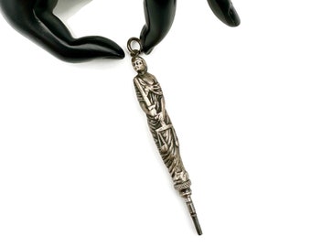 May include: A silver-toned, detailed,  pencil-shaped pendant featuring a figure of Lady Justice holding scales and a sword. The pendant is on a black hand.