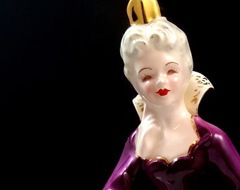 May include: A porcelain figurine of a woman wearing a purple gown with a gold crown. The figurine is standing on a white surface.