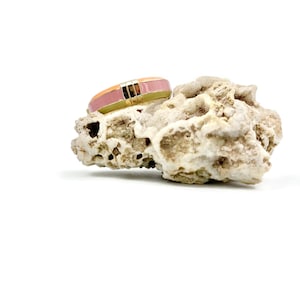 May include: A gold ring with a pink and orange enamel band. The ring is sitting on a piece of white coral.
