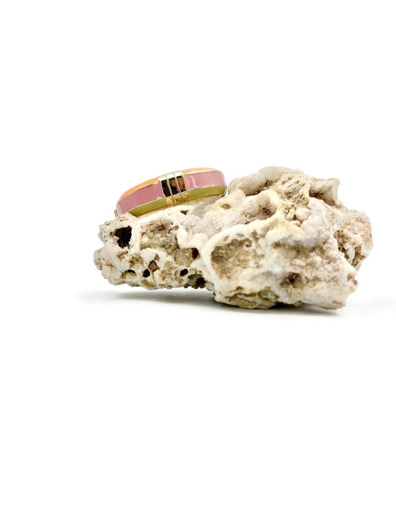 May include: A gold ring with a pink and orange enamel band. The ring is sitting on a piece of white coral.