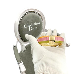 May include: A gold bangle bracelet with a pink, orange, and green enamel design. The bracelet is being held by a gloved hand in front of a small, gray, velvet-covered chair with the words "Christian Dior" printed on the chair.