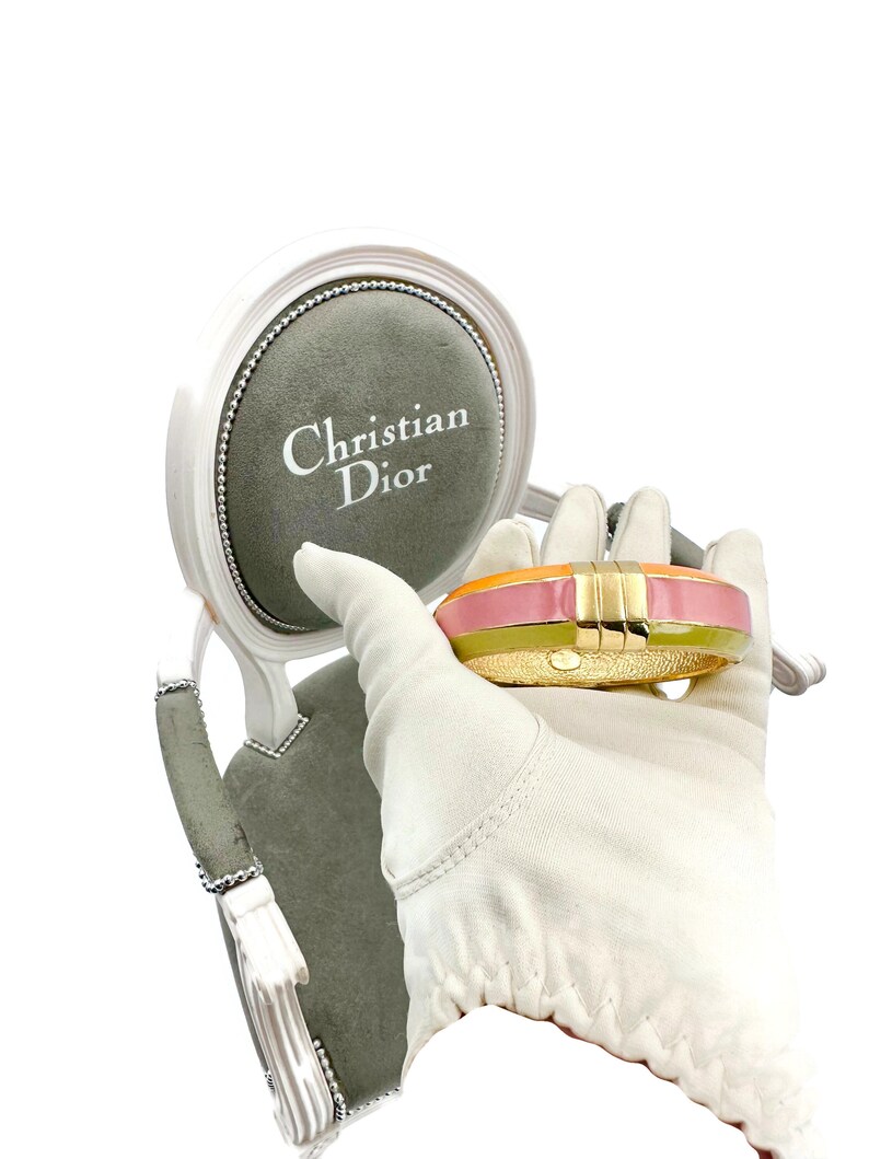 May include: A gold bangle bracelet with a pink, orange, and green enamel design. The bracelet is being held by a gloved hand in front of a small, gray, velvet-covered chair with the words "Christian Dior" printed on the chair.