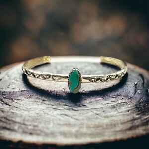 May include: A silver bracelet with a turquoise stone set in the centre. The bracelet has a decorative design of small, half-circle shapes around the edge.