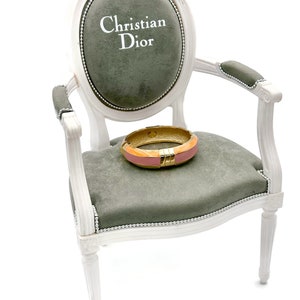 May include: A white armchair with a gray velvet seat and back. The back of the chair has an oval-shaped cushion with the text "Christian Dior" embroidered in white. A pink and gold bangle bracelet is resting on the seat.