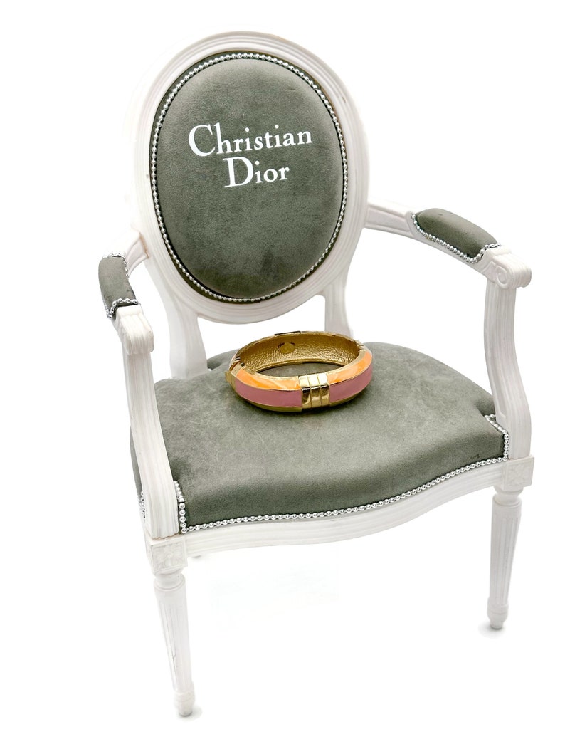 May include: A white armchair with a gray velvet seat and back. The back of the chair has an oval-shaped cushion with the text "Christian Dior" embroidered in white. A pink and gold bangle bracelet is resting on the seat.
