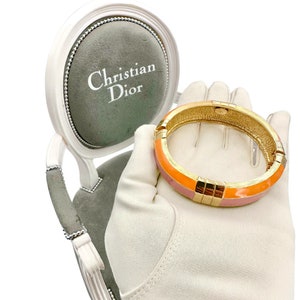 May include: A pink and orange enamel bangle bracelet with gold accents. The bracelet is on a white glove and a small chair with a gray cushion that says "Christian Dior" on it.