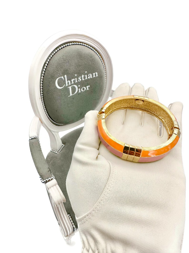 May include: A pink and orange enamel bangle bracelet with gold accents. The bracelet is on a white glove and a small chair with a gray cushion that says "Christian Dior" on it.
