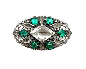 May include: A silver brooch with a large clear rhinestone in the center and smaller green rhinestones around it. The brooch is in the shape of a leaf with a delicate design.