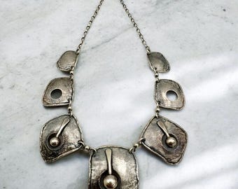 Puede incluir: Un collar de plata con un diseño geométrico. El collar presenta varios colgantes grandes, planos, de plata con bordes redondeados y pequeños círculos cortados en el centro. Cada colgante tiene una pequeña cuenta redonda de plata colgando de él.