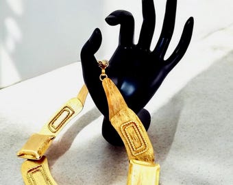 Puede incluir: Un collar dorado de estilo statement con un diseño geométrico. El collar está hecho de piezas rectangulares unidas con una superficie texturizada.