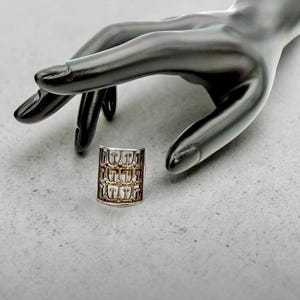 Könnte beinhalten: Ein goldfarbener Ring mit einem sich wiederholenden Muster des Buchstabens "T" wird gezeigt. Der Ring ist zylindrisch und liegt auf einer hellgrauen Oberfläche. Eine schwarze Handmannequin ist im Hintergrund.