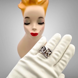 Pode incluir: Um anel de prata com elos de corrente e acabamento texturizado. O anel é usado em uma luva branca.