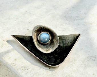 May include: A silver brooch featuring a blue gemstone set within a silver oval. The brooch is shaped like a triangle with rounded edges.
