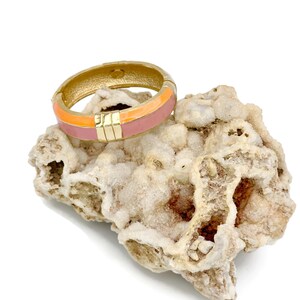 May include: A gold bangle bracelet with a pink and orange enamel design. The bracelet is sitting on a white rock.