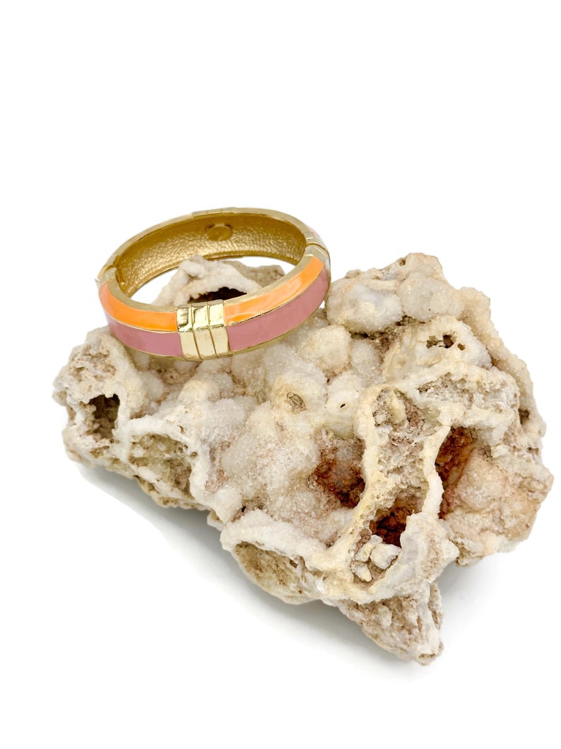 May include: A gold bangle bracelet with a pink and orange enamel design. The bracelet is sitting on a white rock.