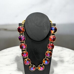 May include: A gold chain necklace featuring large, faceted, octagonal-shaped gemstones. The gemstones are iridescent and display shades of red, pink, and purple.
